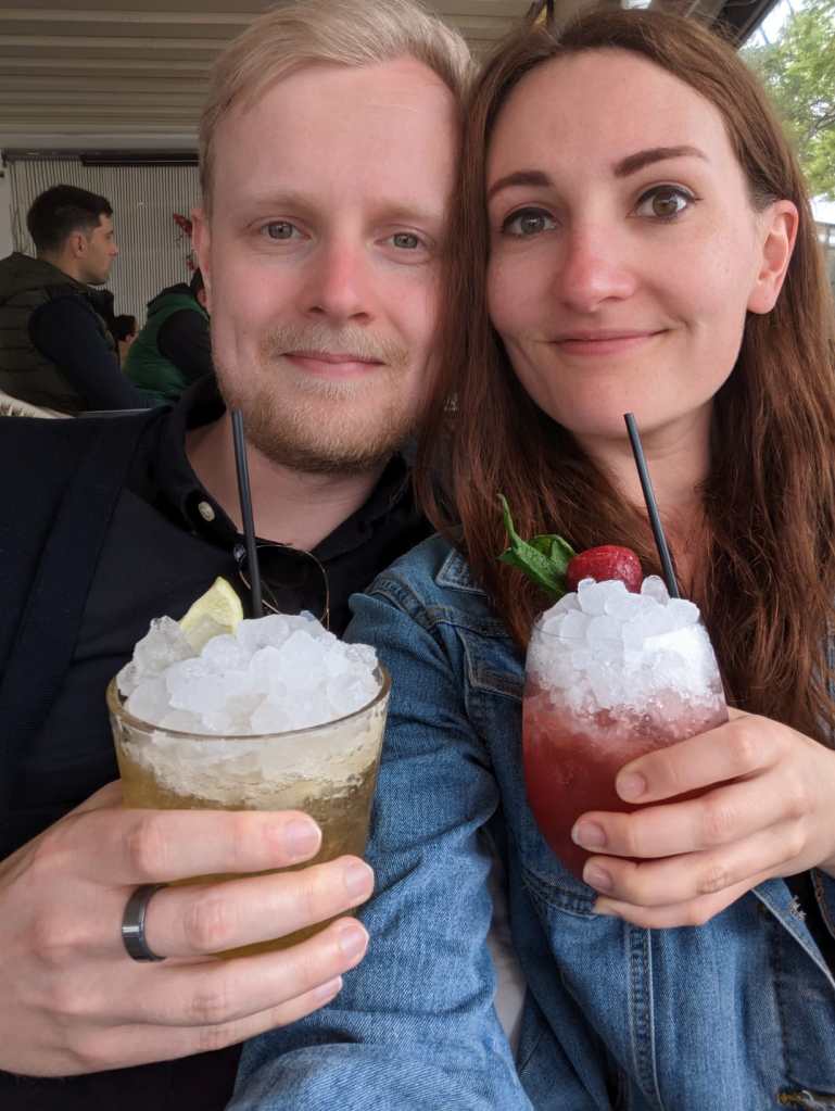 Jake and Claire smiling, holding two cocktails densely packed with ice.